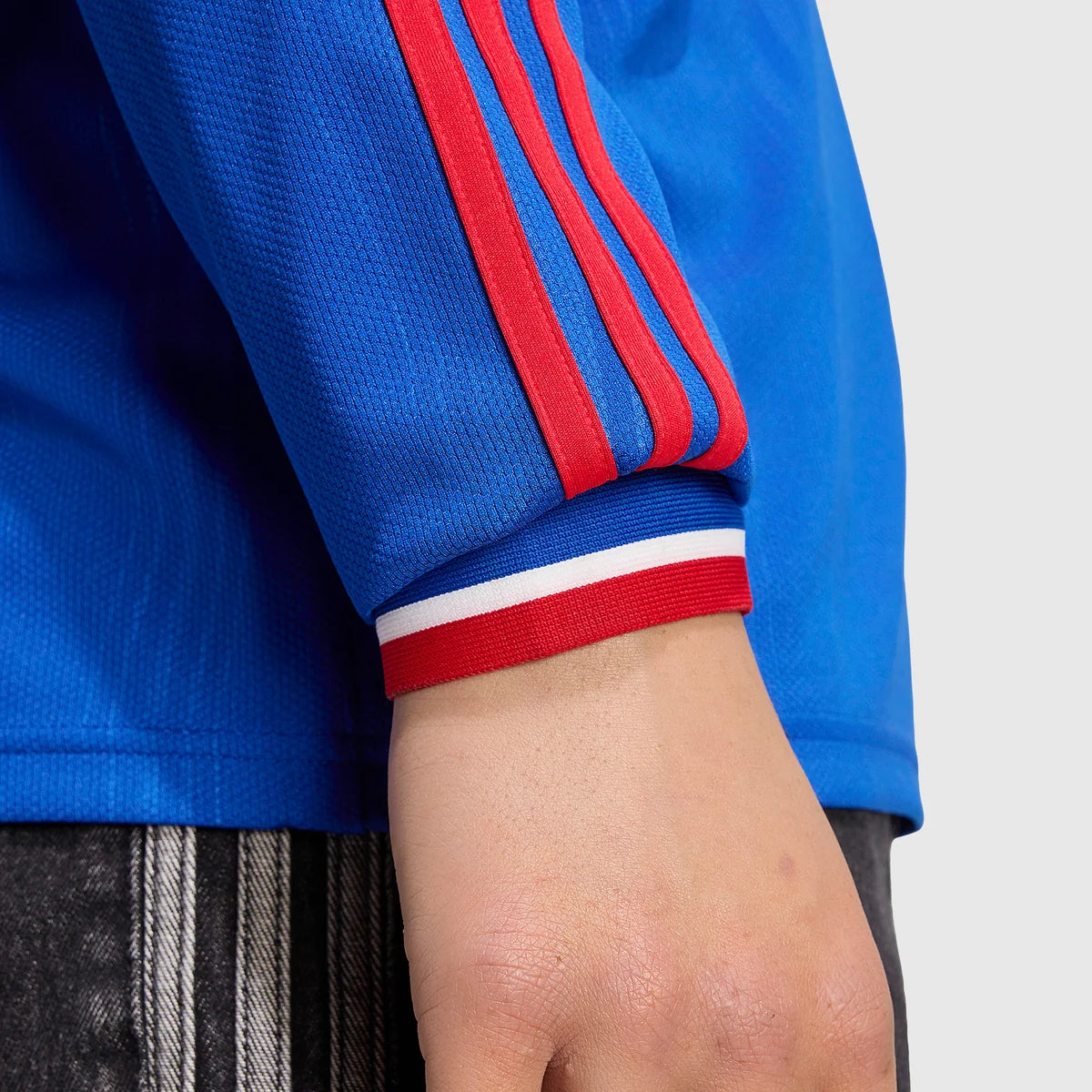 Close-up of a person wearing a bright blue athletic jacket with three red stripes on the sleeve and a red-and-white striped ribbed cuff, shown against a light gray background.