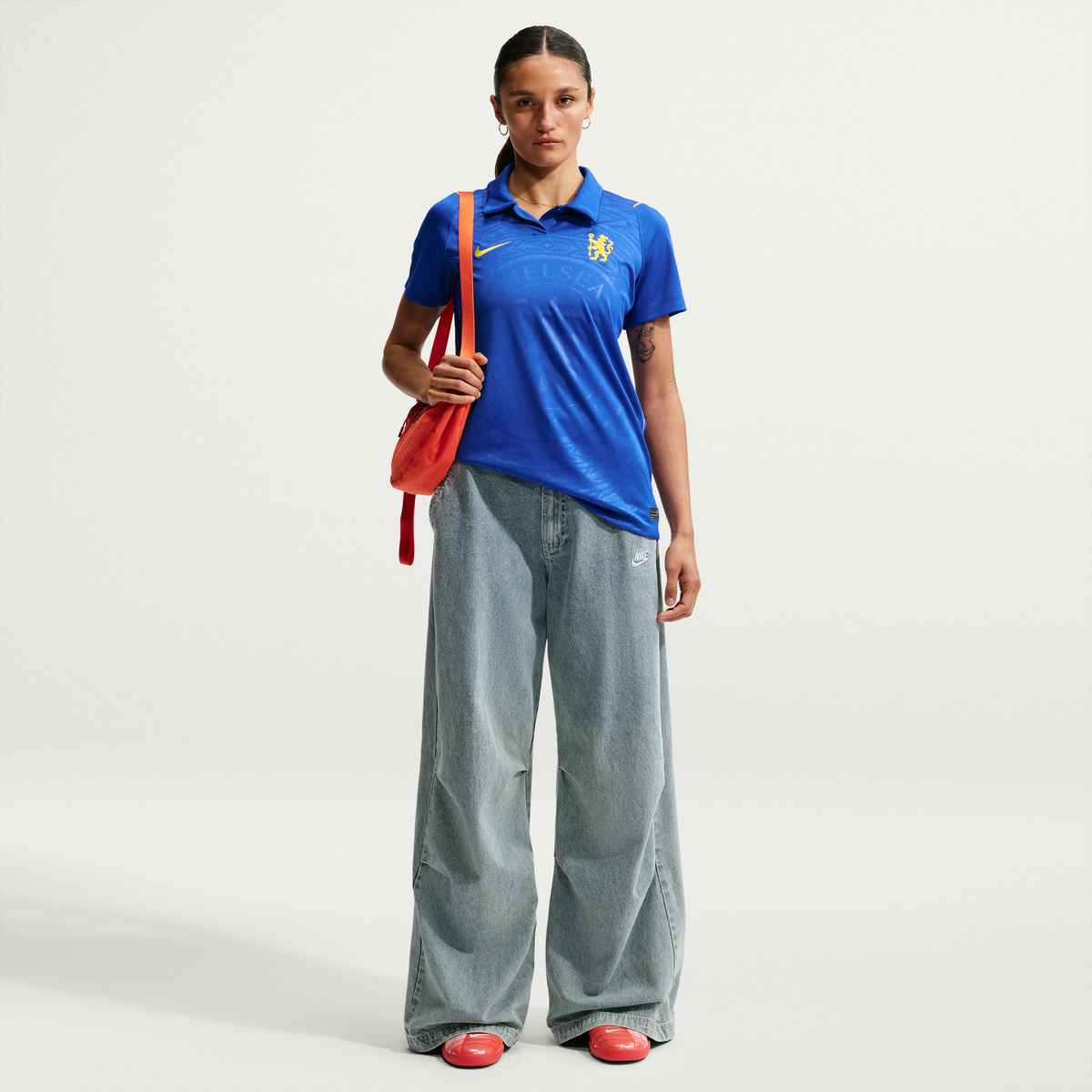Person wearing a blue sports jersey with a logo, holding an orange bag, and wearing red shoes on a white background